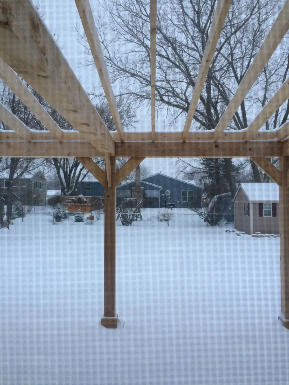 Snow‑covered backyard viewed through a window screen, with a wooden pergola in the foreground and houses, bare trees, and a small shed in the distance under a blanket of spring snow.