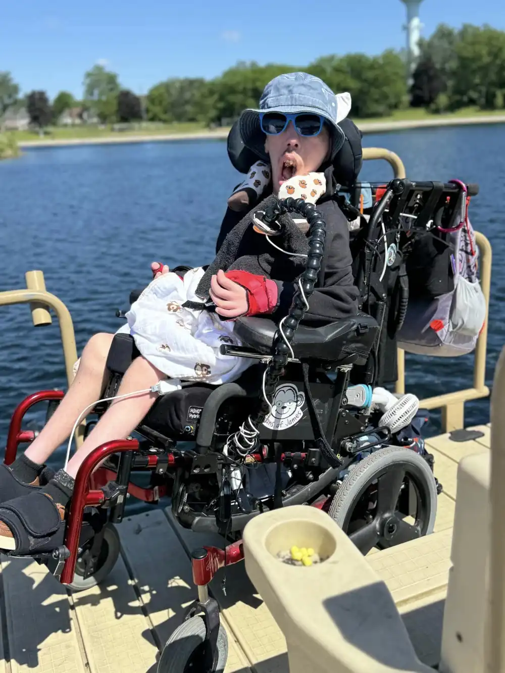 A boy in a wheelchair wearing a bucket hat and sunglasses on a dock overlooking a lake.