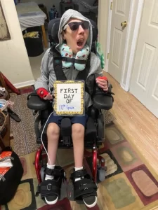 A young boy in a wheelchair wearing a gray hoodie and sunglasses with a sign that reads "First Day of 10th Grade".