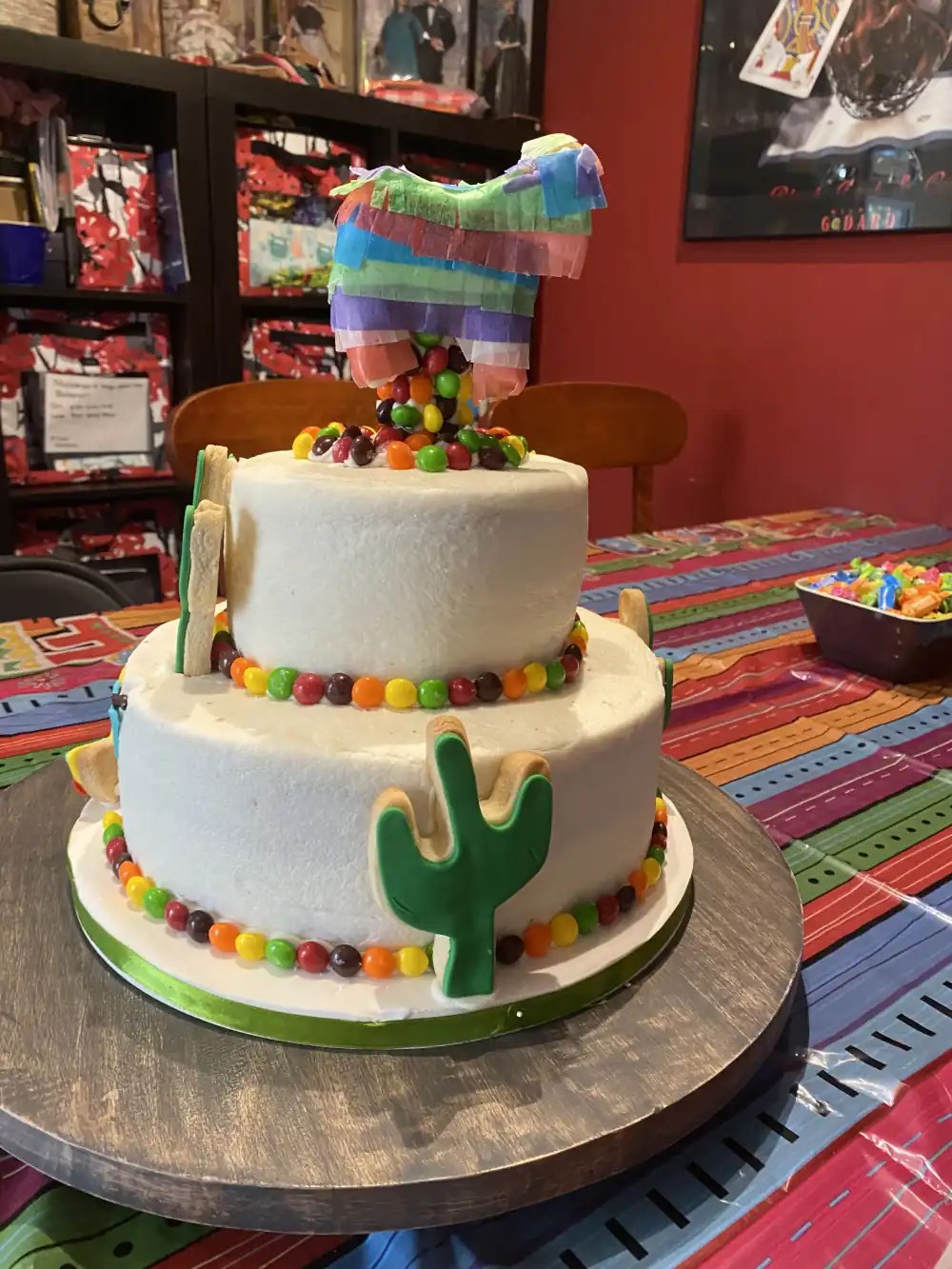 A two-layered white cake decorated with cactus-shaped cookies, Skittles around the lower edge of each layer. There is a donkey-shaped pinata on top of the cake with Skittles coming out of it.