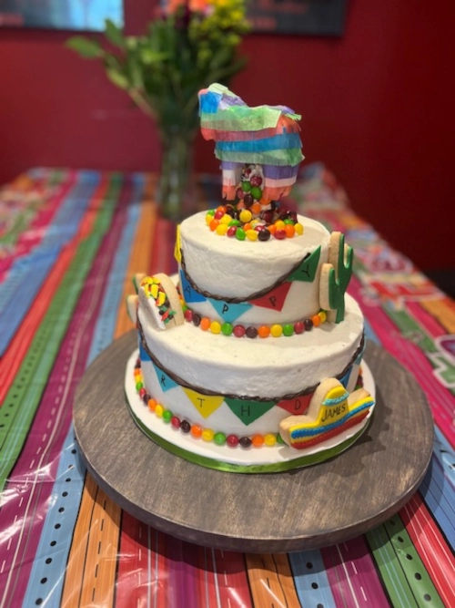 A two-layered white cake decorated with cactus- and sombrero-shaped cookies, and Skittles around the lower edge of each layer. There is a donkey-shaped pinata on top of the cake with Skittles coming out of it.