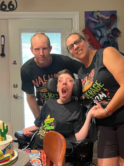 A man and a woman smiling as they stand behind a boy in a wheelchair.